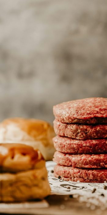 Stack of raw beef patties on a rustic table with buns, ideal for burger preparation theme.